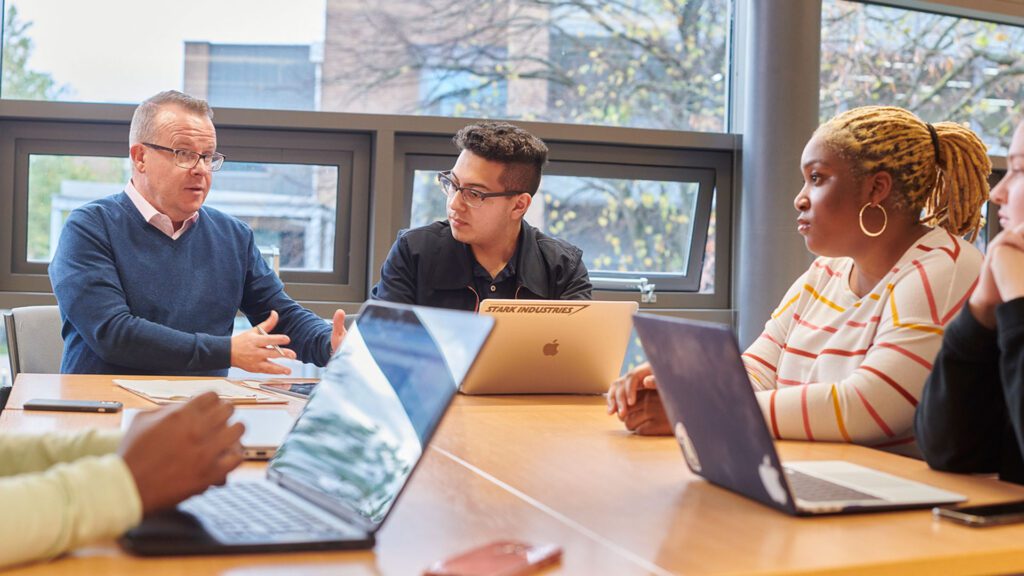 A group of students sat with an academic around a table. Each student has their own laptop.